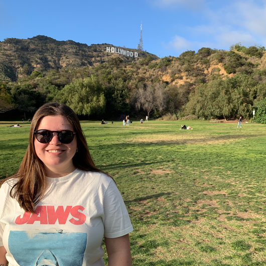 Female, early 20s, with brown hair and wearing sunglasses, standing in a green open field with the Hollywood sign on the hill behind